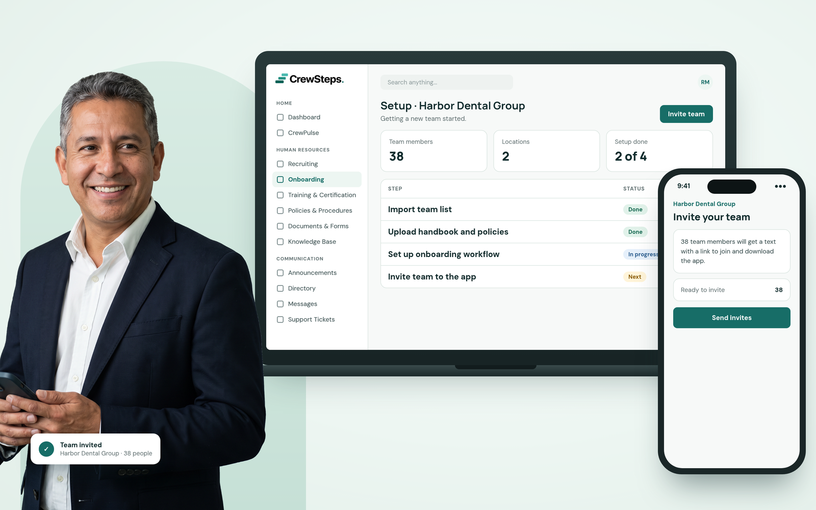 A confident business consultant in a navy blazer holding his phone beside a CrewSteps laptop showing setup steps for a new client team and a phone ready to invite 38 team members.