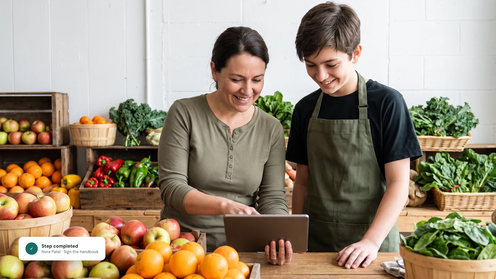 A grocery store owner and a young clerk in an apron smile at a tablet together behind a produce counter.